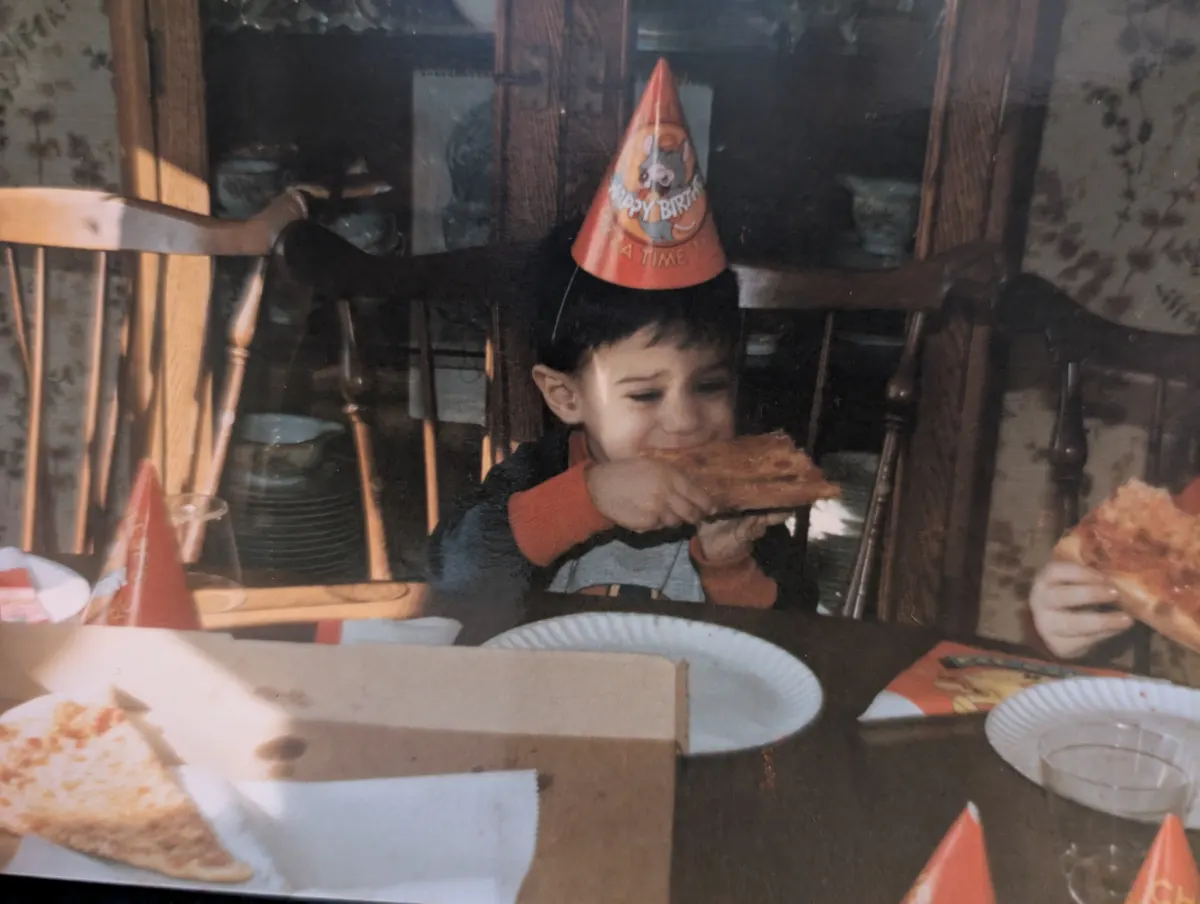 The site's founder as a young boy in a paper party hat, biting into a slice at a birthday party, with a pizza box and paper plates on the table