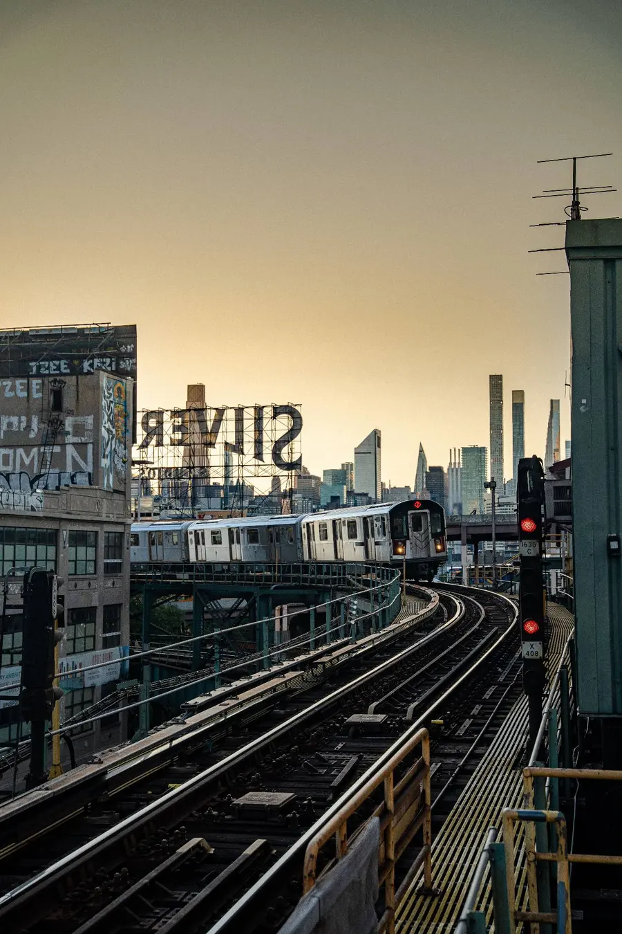 A 7 train rounding the elevated curve in Long Island City at dusk, with the Silvercup sign and the Manhattan skyline behind it