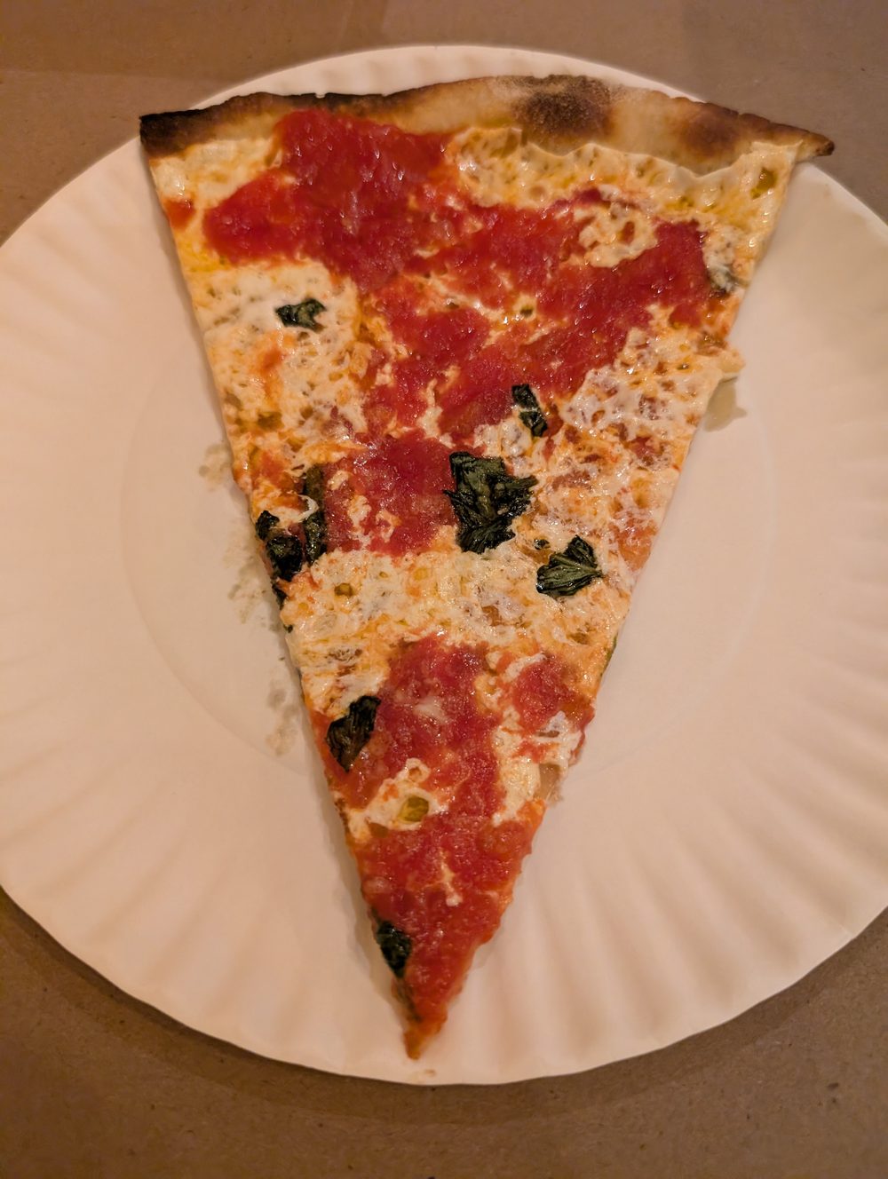 A margherita slice on a paper plate, thin crust charred at the rim, with torn basil across the cheese.