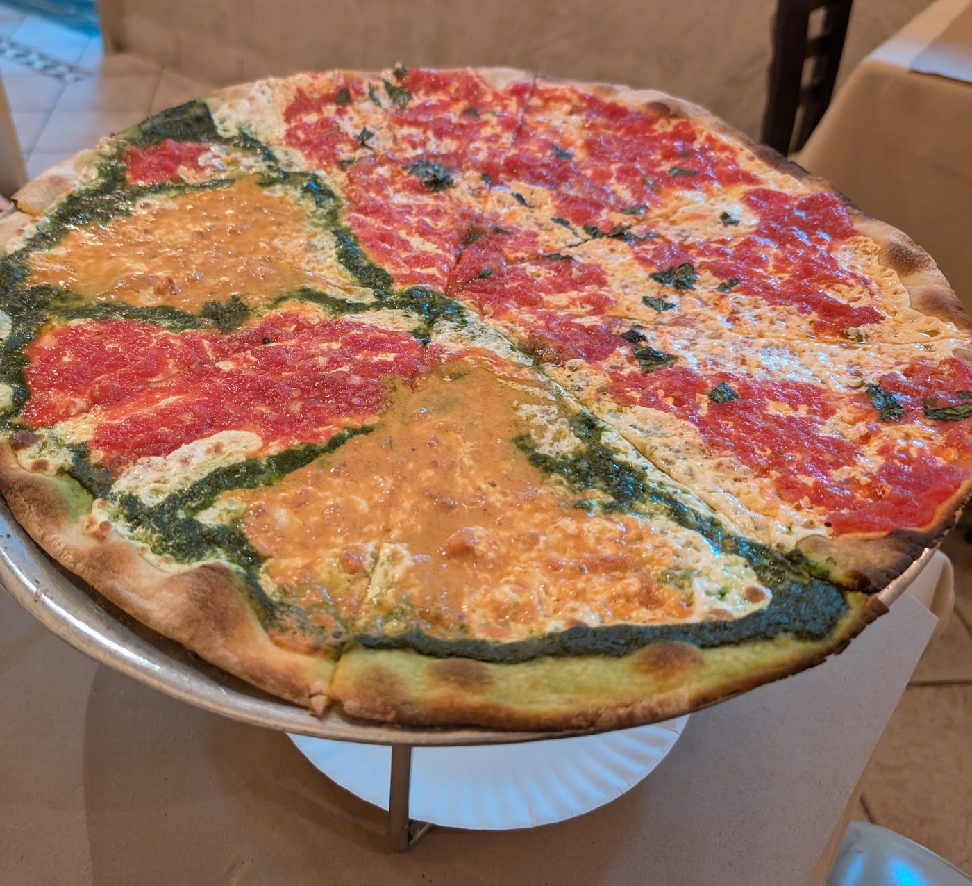 A whole thin-crust pie on a raised metal stand, half of it tri-color with green pesto dividing red marinara and creamy vodka sections, the other half margherita with basil.