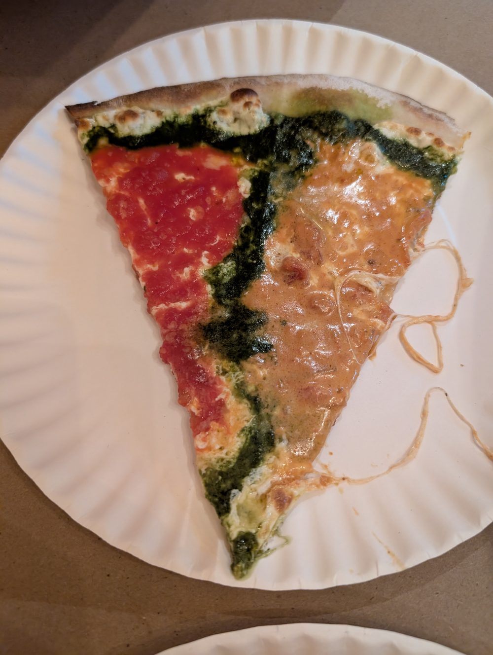 A tri-color slice on a paper plate: marinara on one side, a stripe of pesto down the middle, creamy vodka sauce on the other.