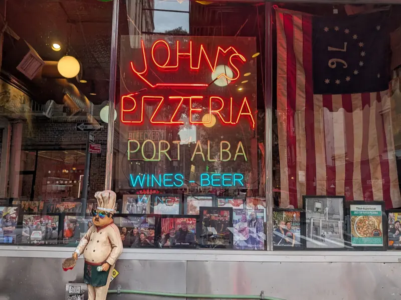 The front window at John's of Bleecker Street: red neon John's Pizzeria and Port'Alba signs over a row of framed photos of famous customers