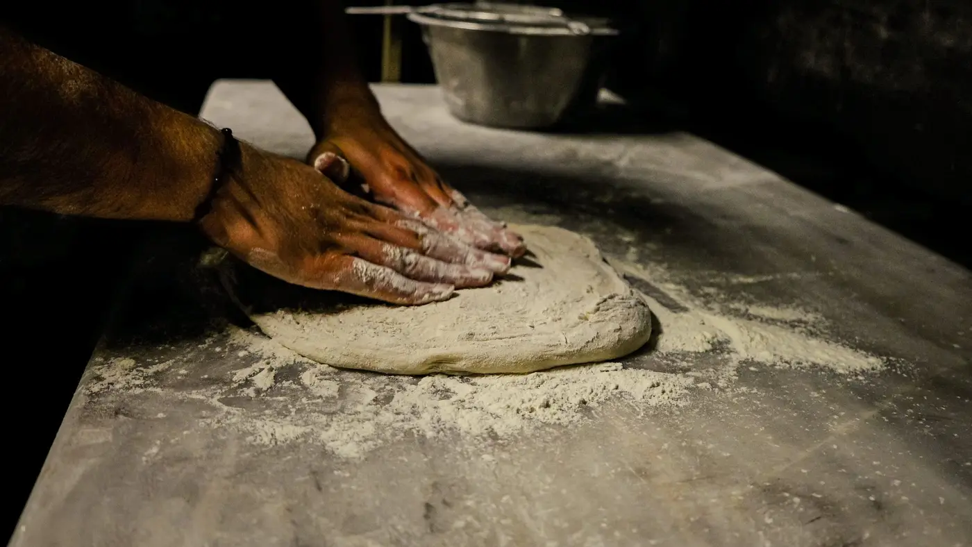 Floured hands pressing out a ball of pizza dough on a marble counter dusted with flour