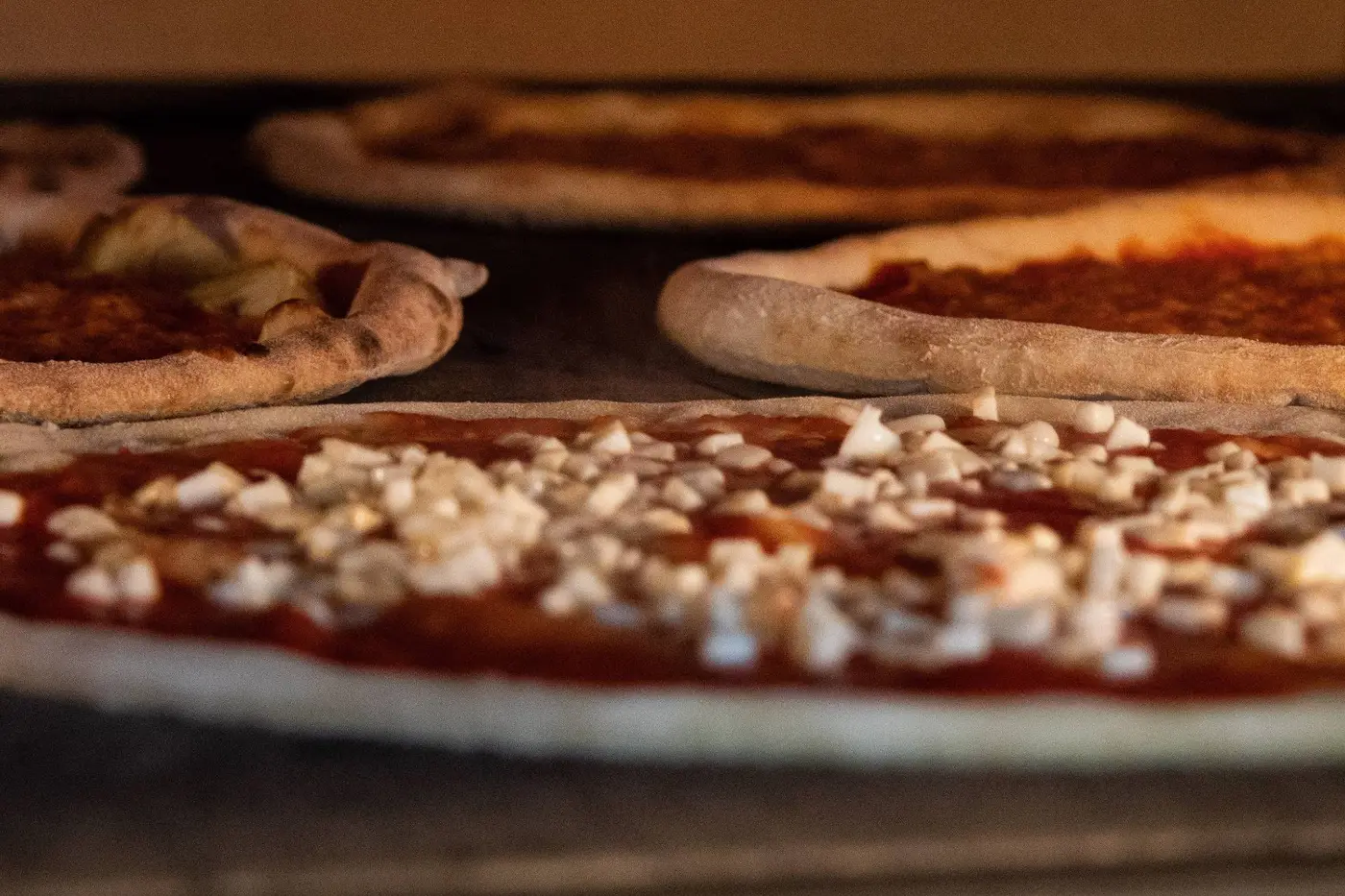 Pies baking side by side in a pizza oven, the nearest one topped with tomato and diced mozzarella