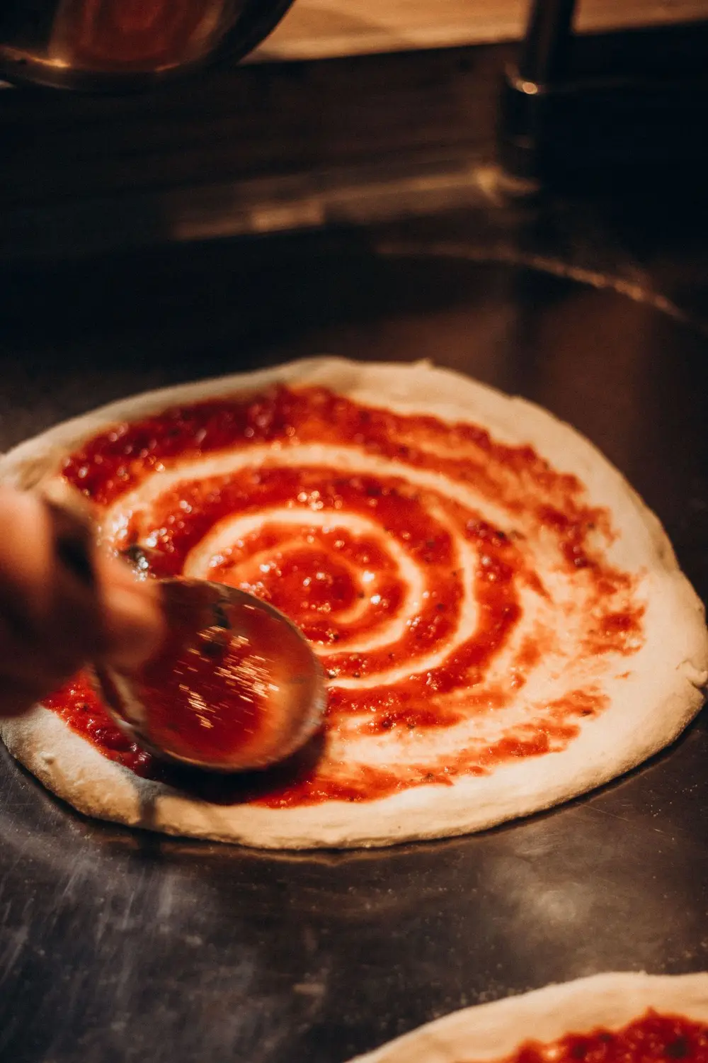 A ladle spiraling tomato sauce out from the center of a stretched pizza skin