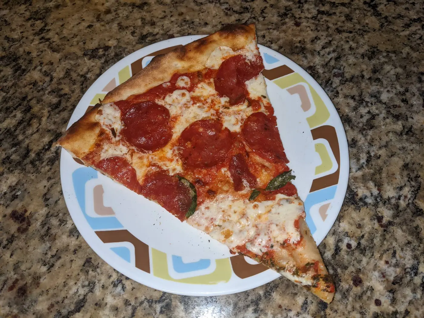 A New York slice with pepperoni and a little basil on a patterned plate, the rim browned and blistered