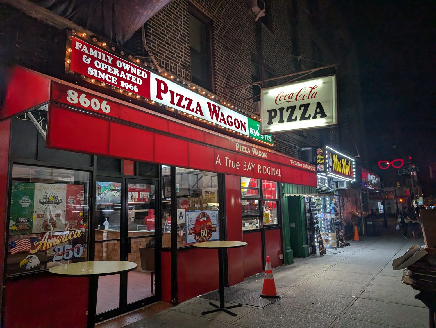 Pizza Wagon at night on 5th Avenue, its marquee lit with bulbs reading Family Owned & Operated Since 1966, with sidewalk tables out front.