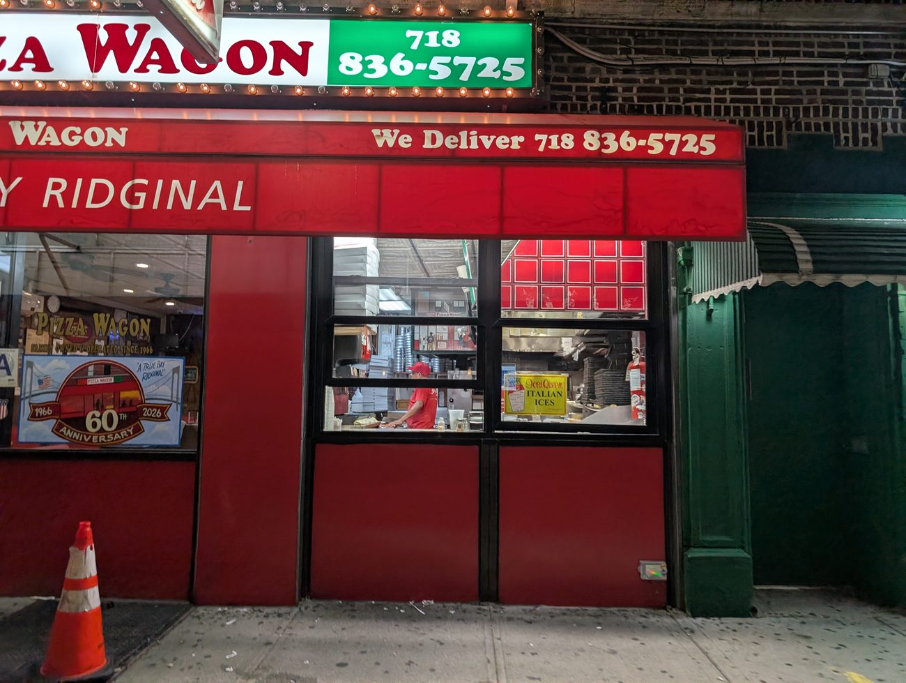 The Pizza Wagon service window open onto the sidewalk, a worker in a red cap inside, with stacked pizza boxes and an Italian ices sign.
